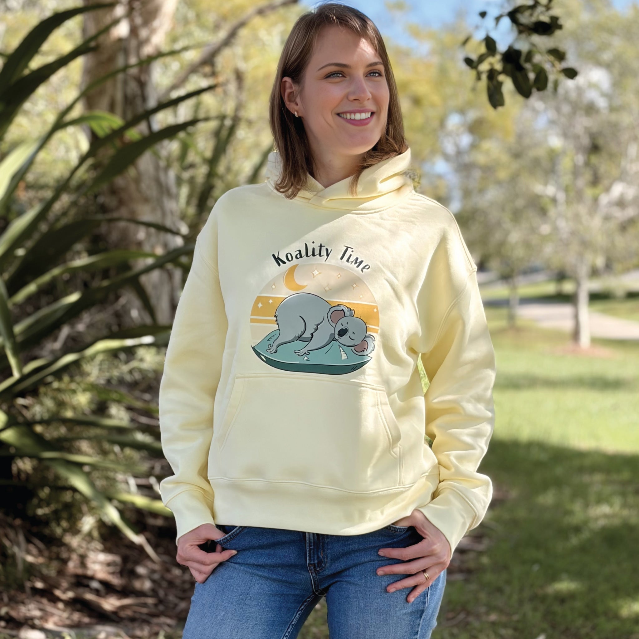 Woman wearing a Butter Women's Koala Hoodie featuring a hand-drawn koala illustration and "Koality Time" slogan, enjoying a relaxed outdoor lifestyle setting in a cosy and comfortable hoodie.