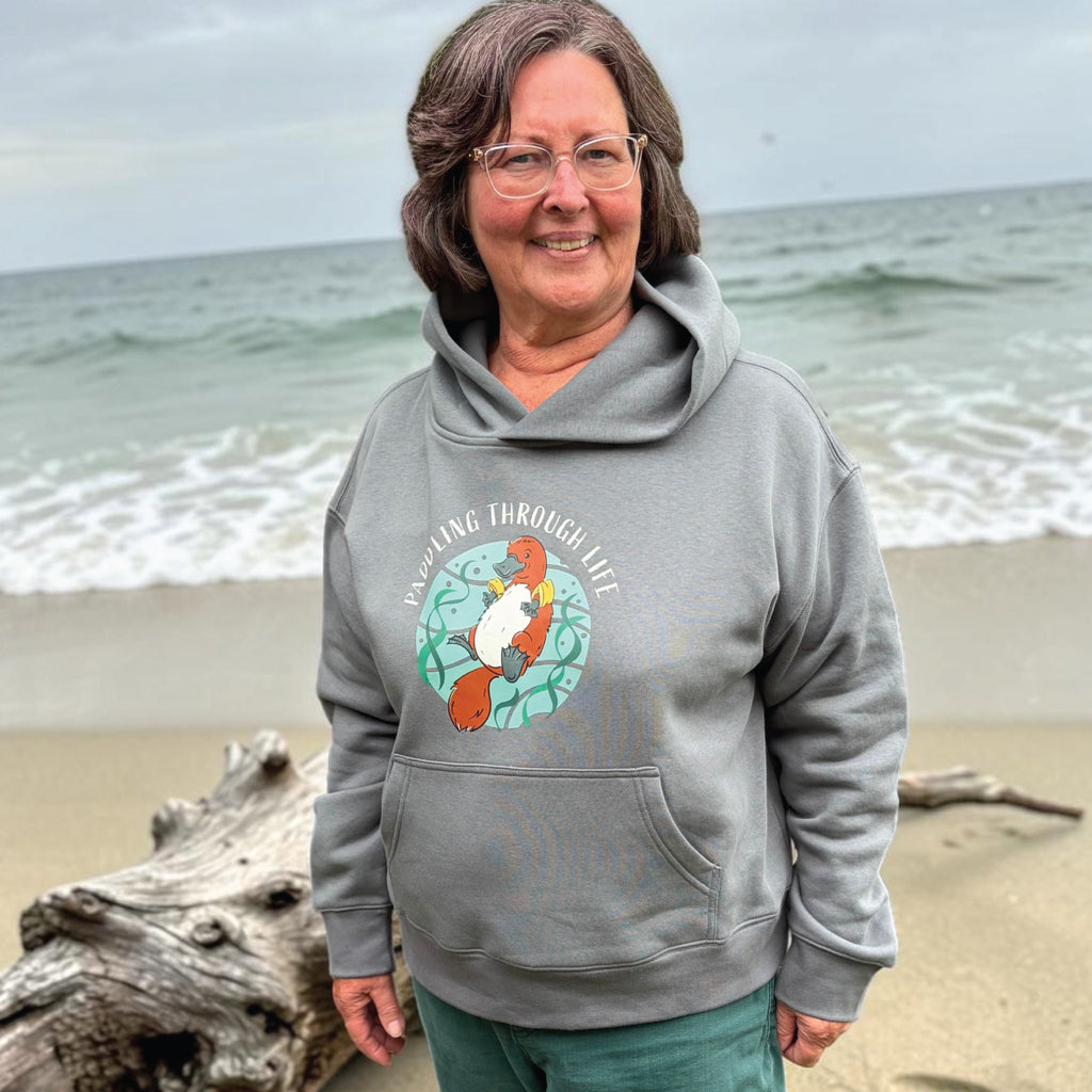 Woman wearing a Funny Platypus Hoodie featuring a hand-drawn Australian platypus design and "Paddling Through Life" slogan, styled in a relaxed outdoor lifestyle setting.