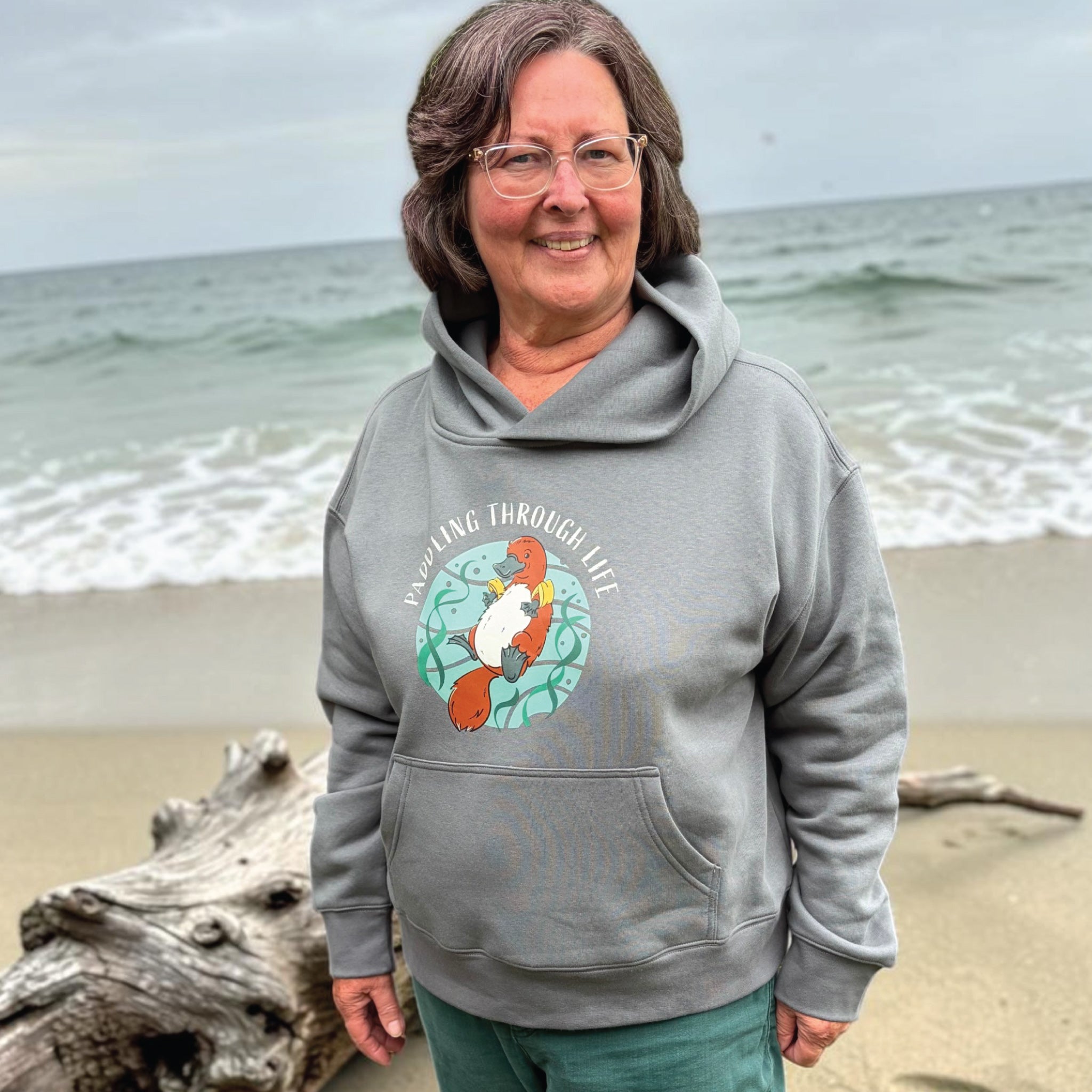 Woman wearing a Funny Platypus Hoodie featuring a hand-drawn Australian platypus design and "Paddling Through Life" slogan, styled in a relaxed outdoor lifestyle setting.