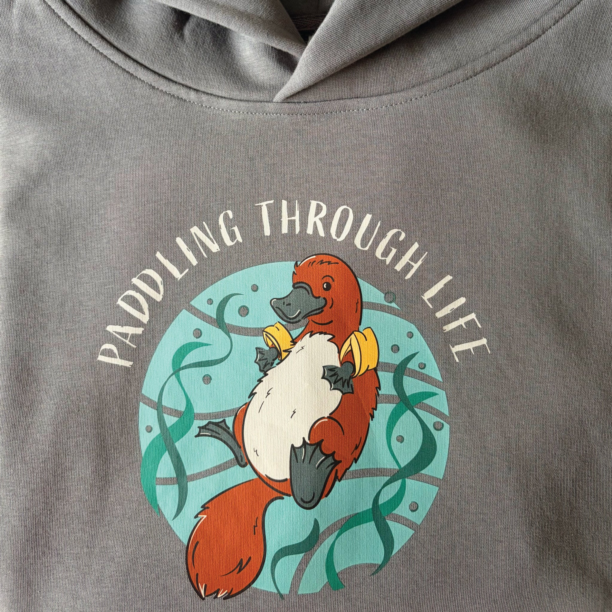 Funny Platypus Women's Hoodie featuring a hand-drawn Australian platypus design and "Paddling Through Life" slogan, styled in a relaxed outdoor lifestyle setting.