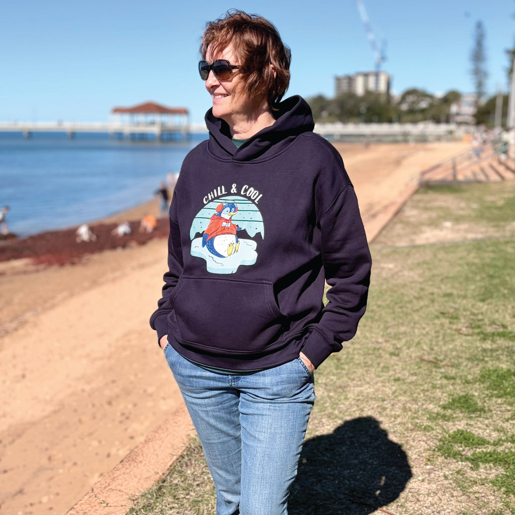 Woman wearing a cosy Penguin Ink Hoodie featuring a hand-drawn penguin illustration and "Chill & Cool" slogan, styled in a relaxed outdoor lifestyle setting.