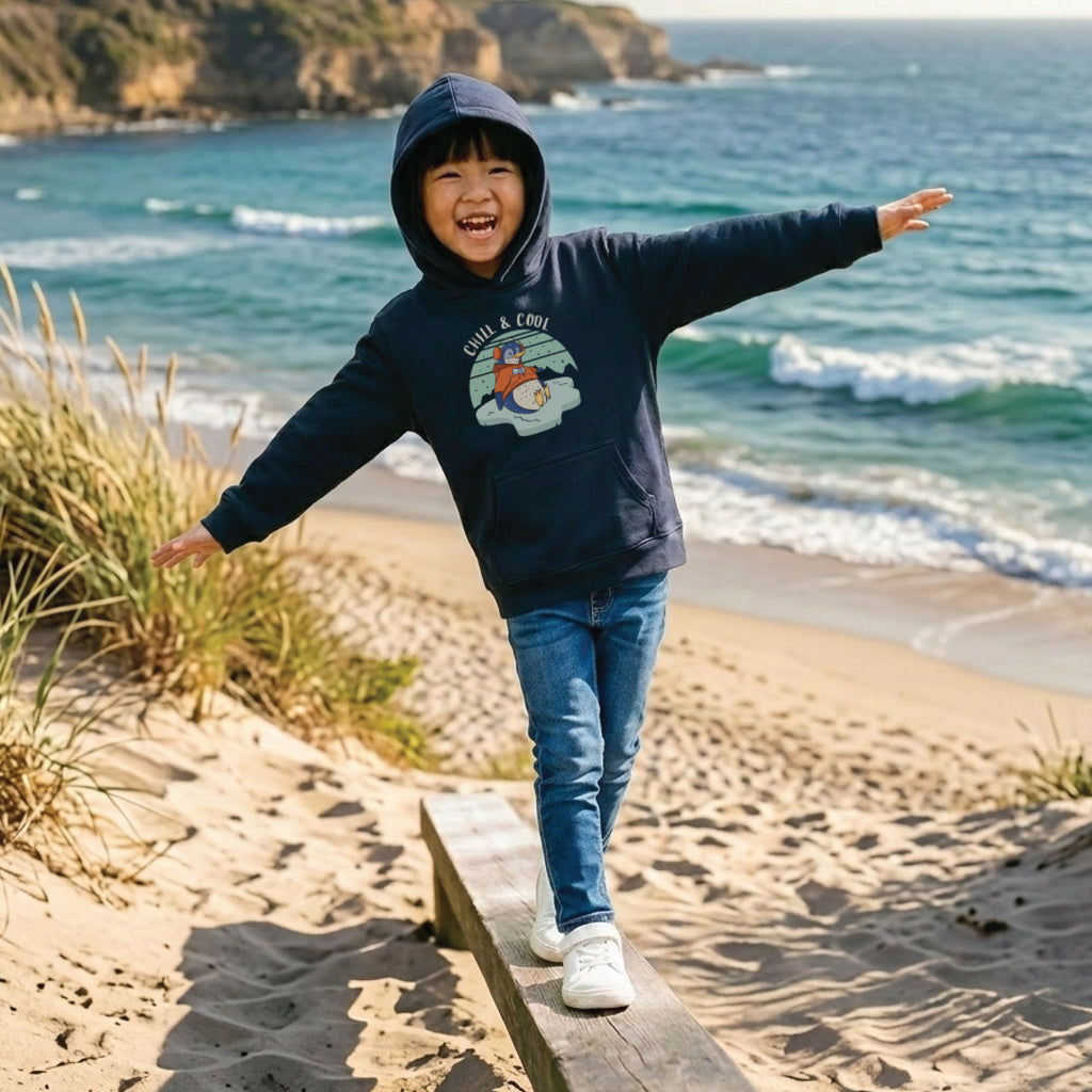 Child wearing a Kids' Penguin Hoodie featuring a hand-drawn penguin illustration and "Chill & Cool" slogan, enjoying a fun and relaxed outdoor lifestyle setting.