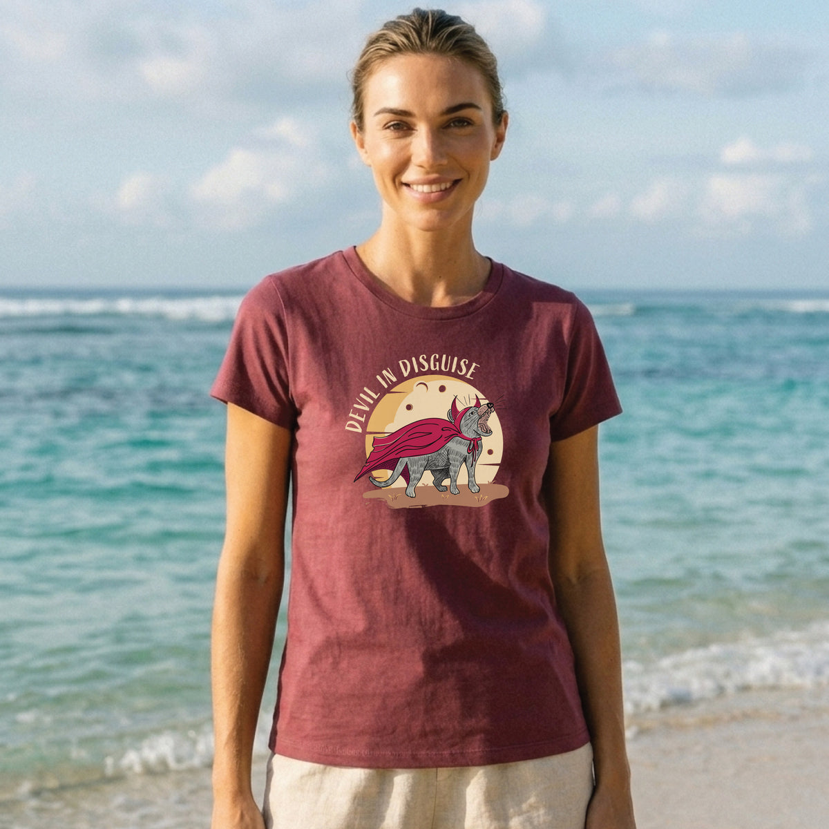 A woman standing on a beach, wearing a berry-coloured T-shirt featuring a hand-drawn graphic of a Tasmanian devil with the text “DEVIL IN DISGUISE.”