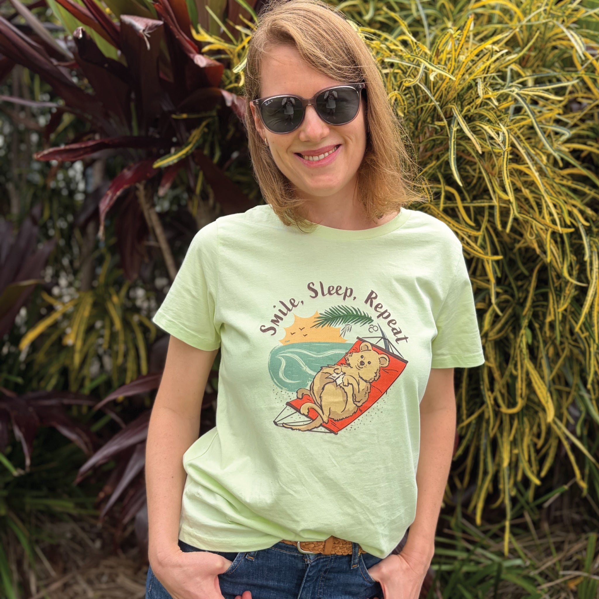 Woman wearing a Lime Women's Quokka T-Shirt featuring a hand-drawn quokka illustration and "Smile, Sleep, Repeat" slogan, enjoying a relaxed outdoor lifestyle setting.