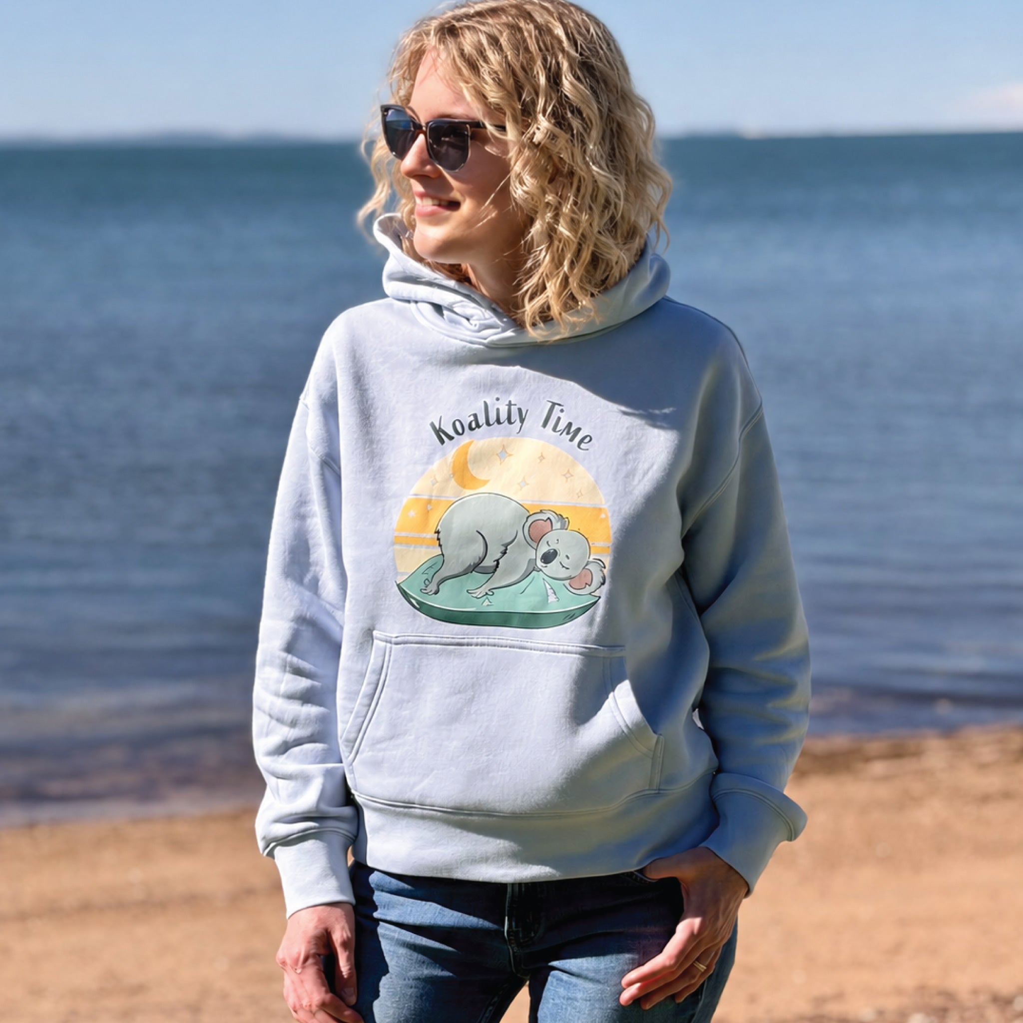Woman wearing a Powder Women's Koala Hoodie featuring a hand-drawn koala illustration and "Koality Time" slogan, enjoying a relaxed outdoor lifestyle setting in a cosy and comfortable hoodie.