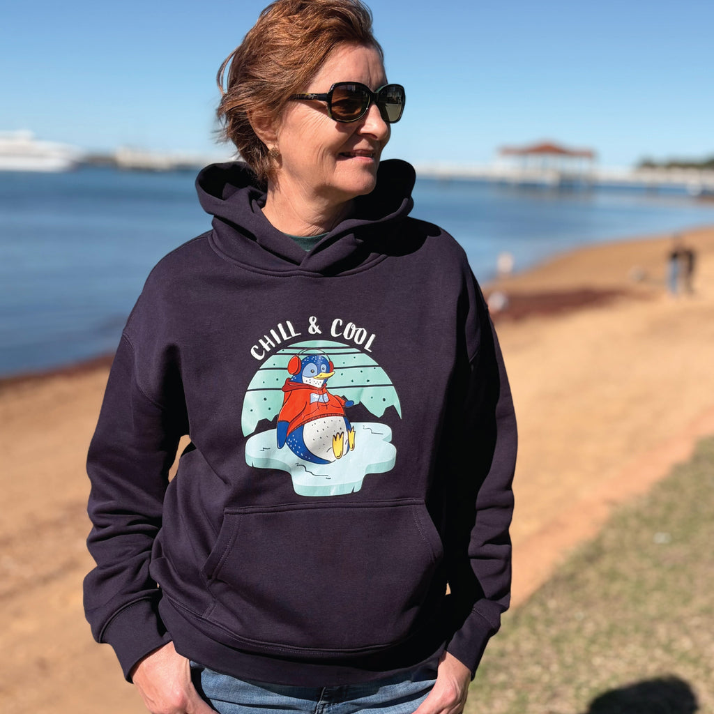 Woman wearing a cosy Penguin Hoodie featuring a hand-drawn penguin illustration and "Chill & Cool" slogan, styled in a relaxed outdoor lifestyle setting.