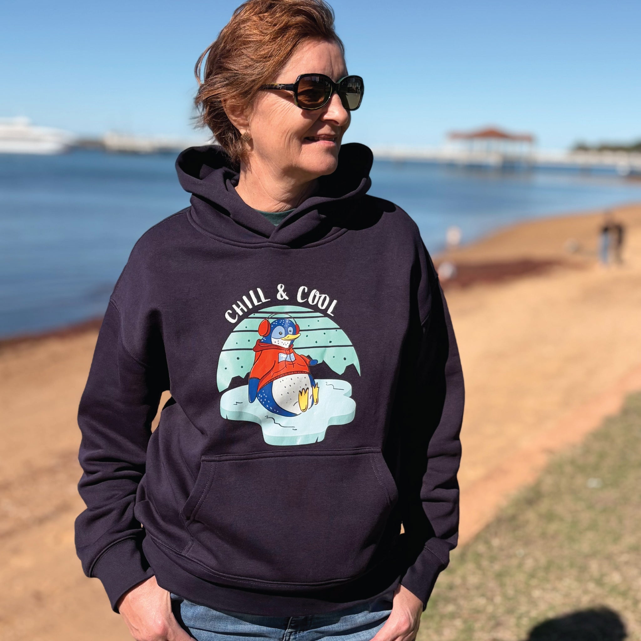Woman wearing a cosy Penguin Hoodie featuring a hand-drawn penguin illustration and "Chill & Cool" slogan, styled in a relaxed outdoor lifestyle setting.