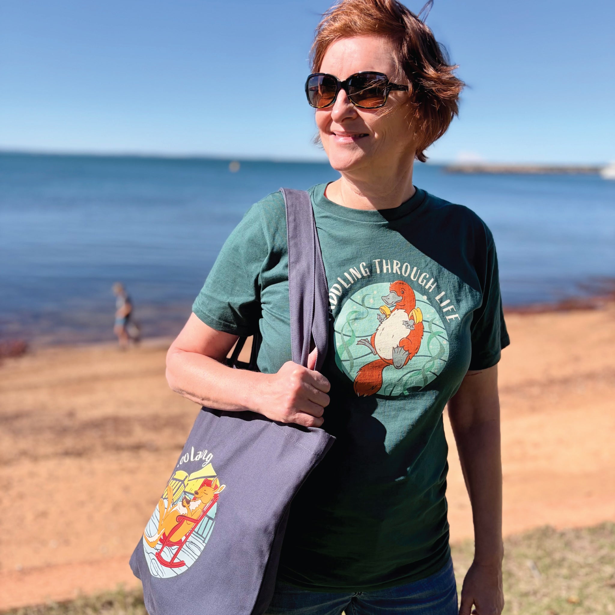 Woman wearing a Women's Platypus T-Shirt featuring a hand-drawn platypus illustration and "Paddling Through Life" slogan, enjoying a relaxed outdoor lifestyle setting.